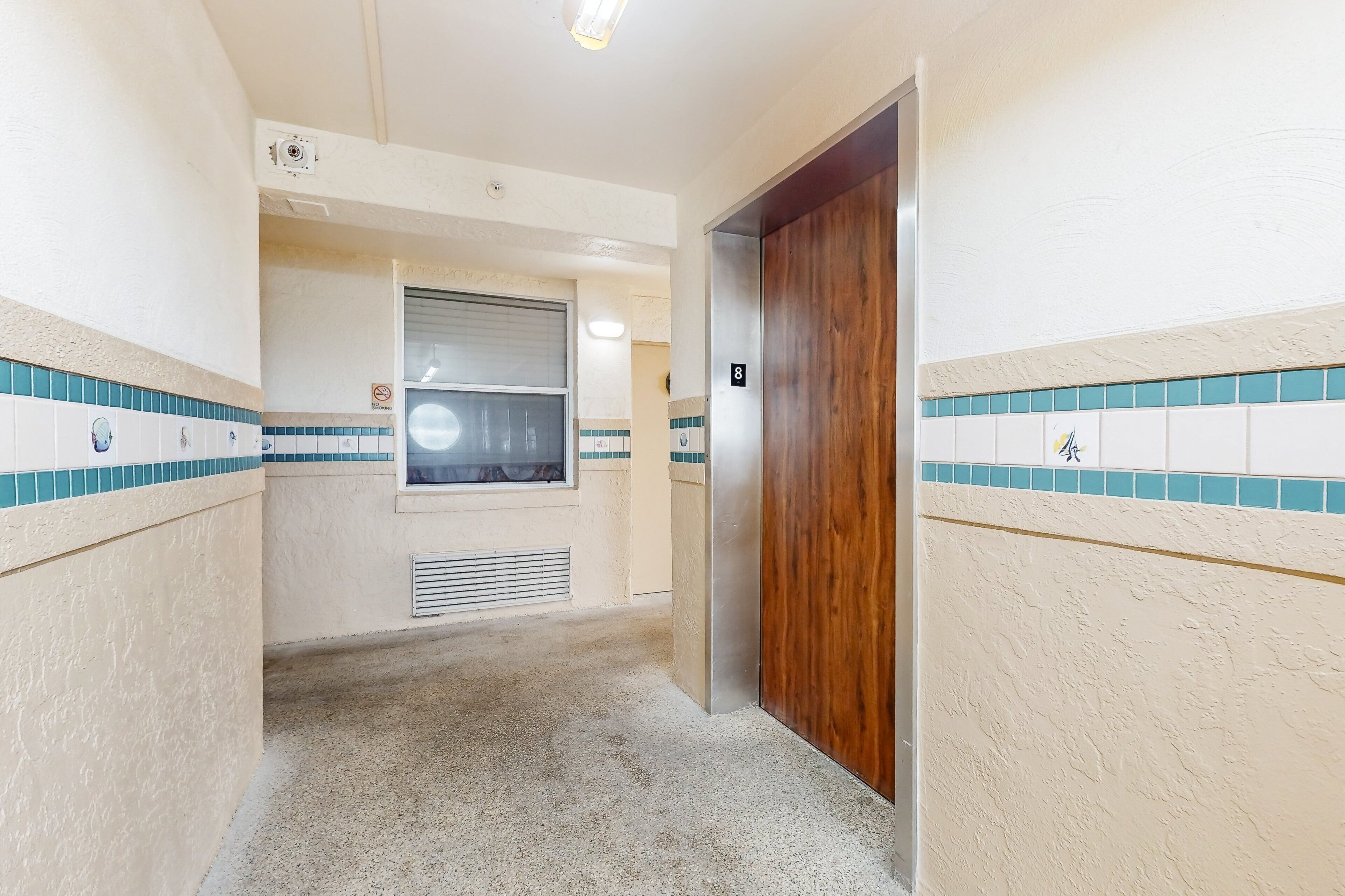 Un couloir étroit avec une porte en bois, une fenêtre avec des stores et un mur carrelé orné d’une bordure bleue.