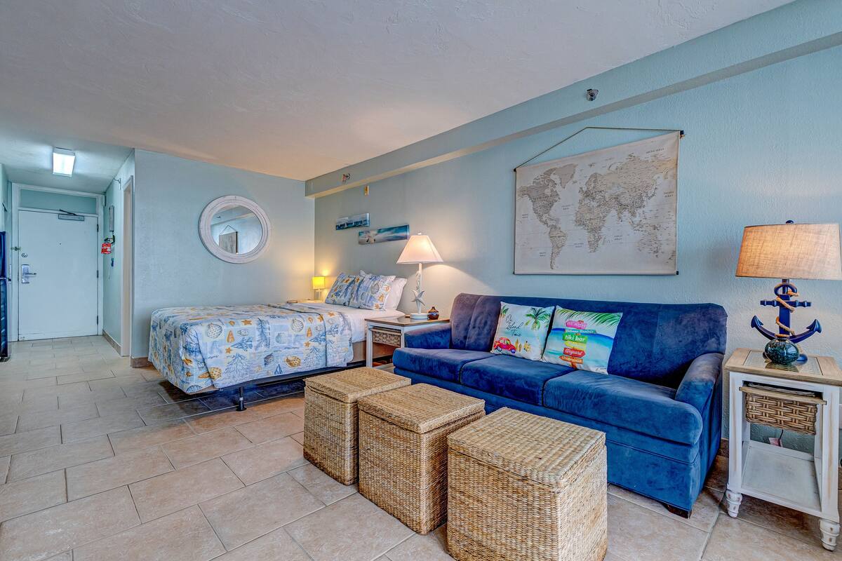 Une pièce comprenant un lit, un canapé bleu, des poufs en osier, une carte accrochée au mur et une lampe posée sur une table d’appoint.