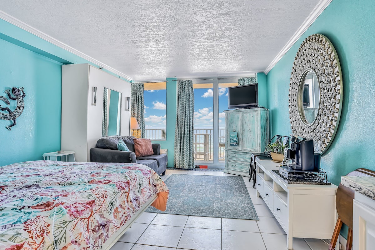 Un dormitorio con cama, cómoda, televisor y vista al cielo a través de la puerta.