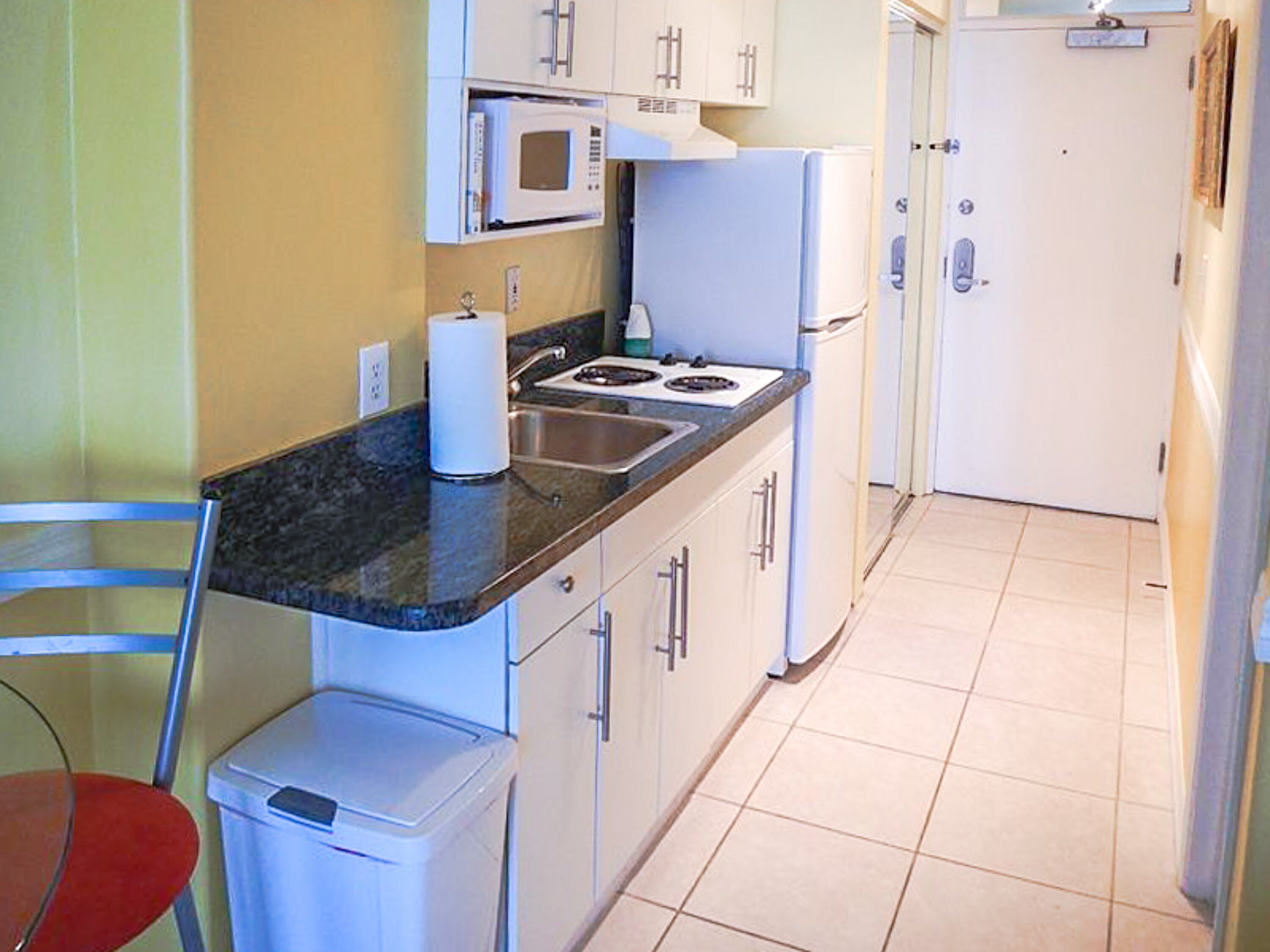 A compact kitchen with a microwave, stove, and refrigerator. There is a trash bin and a chair.