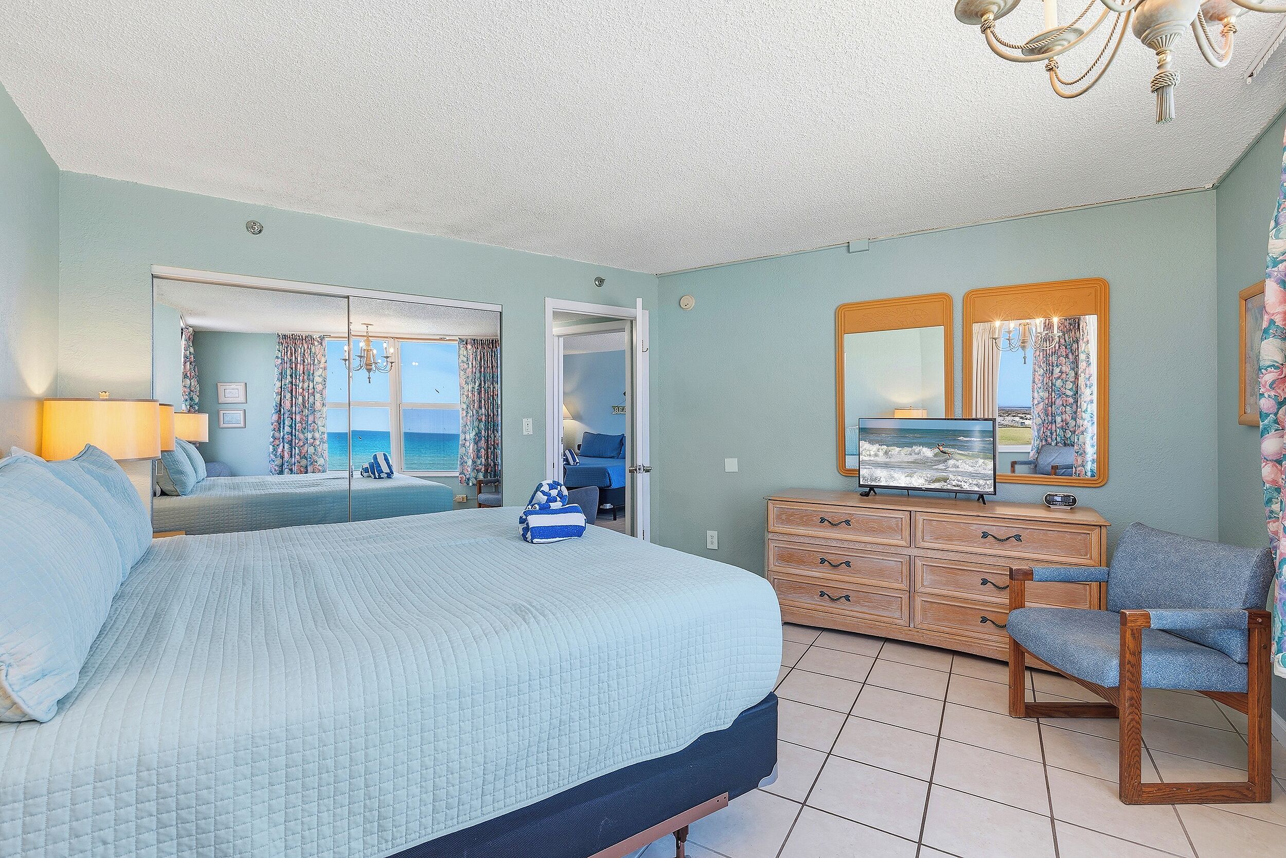A bedroom with a bed, a dresser, a chair, a television, and a view of the sea through a window.