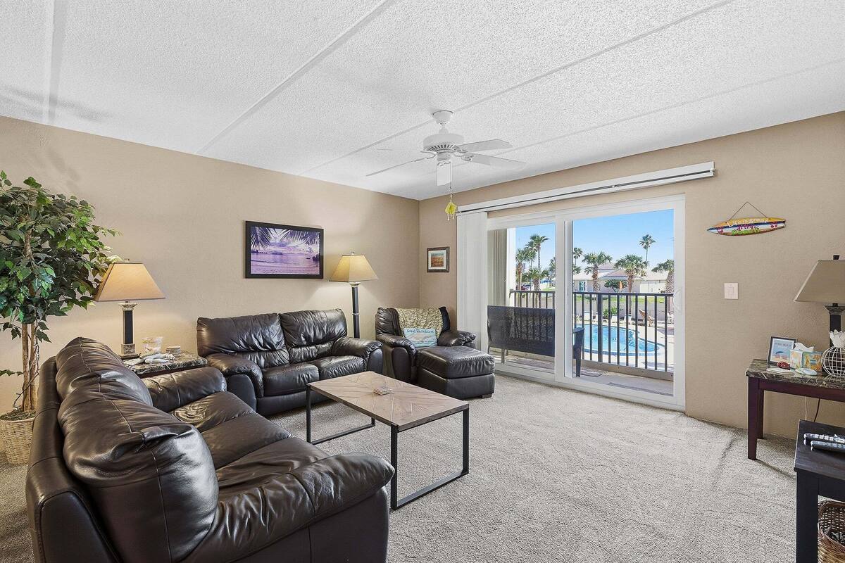 A living room with a sectional sofa, a coffee table, a ceiling fan, and a view of a pool through a sliding glass door.