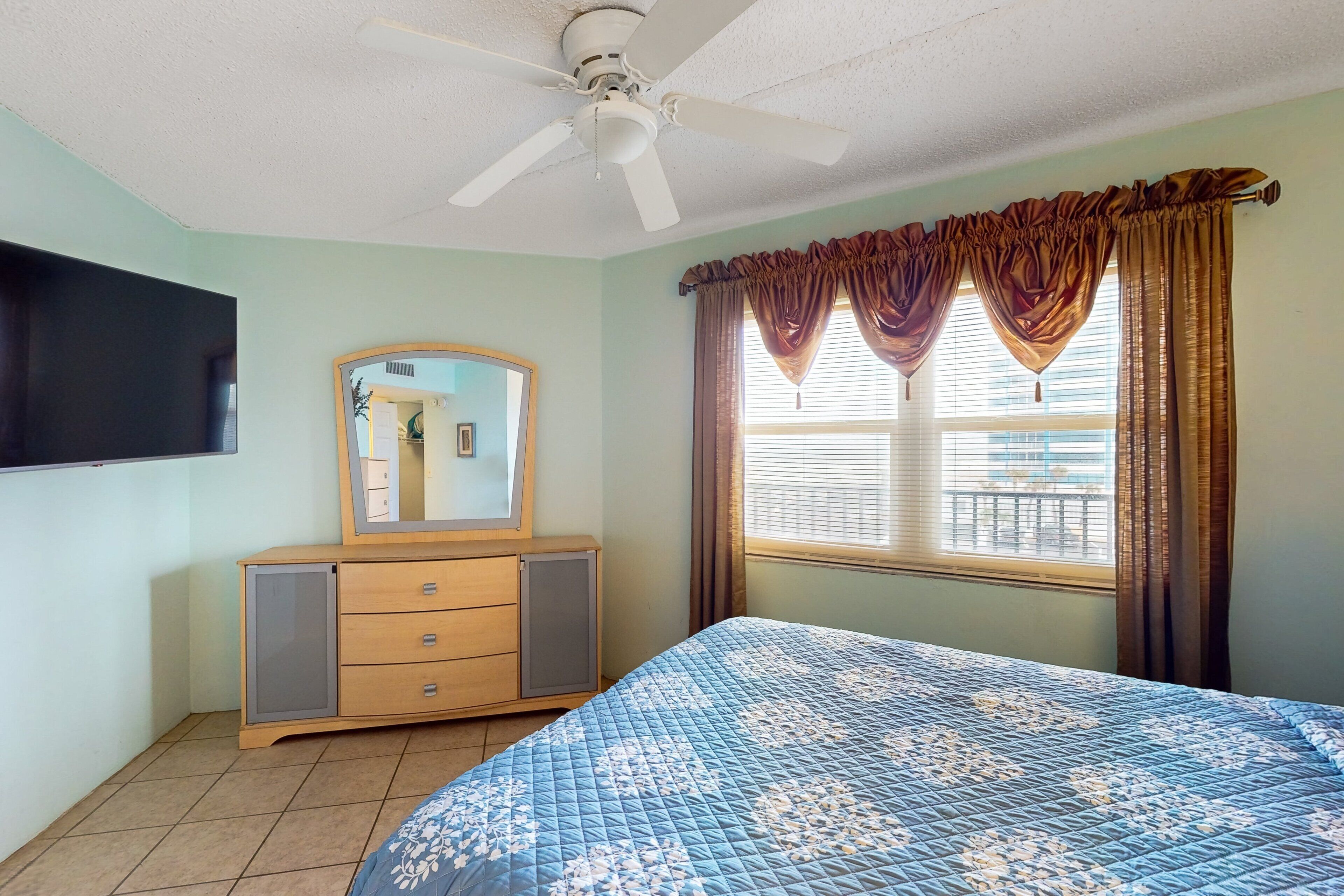 A bedroom with a bed, a dresser, a ceiling fan, a window with curtains, and a flat-screen TV mounted on the wall.