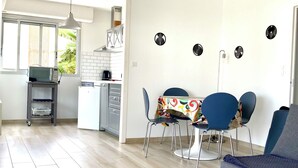 Una cucina moderna con un forno a microonde su uno scaffale, un'area pranzo con sedie blu e una tovaglia colorata, e un divano grigio in un soggiorno luminoso con pavimento in legno.