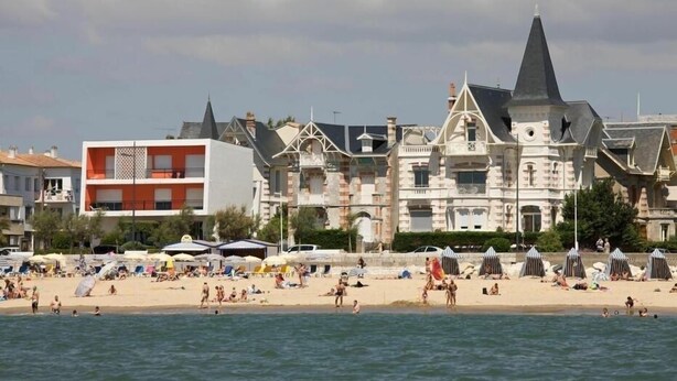 Una spiaggia con persone che nuotano e si rilassano al sole, e una fila di edifici con stili architettonici diversi.