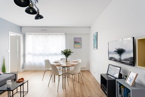 Un salon moderne comprenant un coin repas, un parquet, un canapé gris, une table basse et un téléviseur posé sur un meuble.