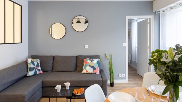 Uma sala de estar moderna com um sofá cinzento, dois espelhos redondos, uma mesa de centro e uma área de jantar com uma cadeira e mesa brancas.