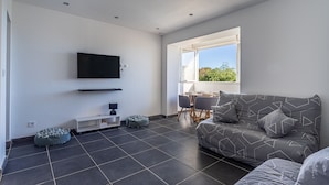 Un salon moderne avec un canapé gris, un téléviseur à écran plat, un coin repas comprenant une table et des chaises, et une grande fenêtre donnant sur un paysage verdoyant.
