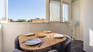 Un coin repas avec une table en bois dressée pour quatre personnes, un ventilateur sur pied et une fenêtre avec des rideaux.