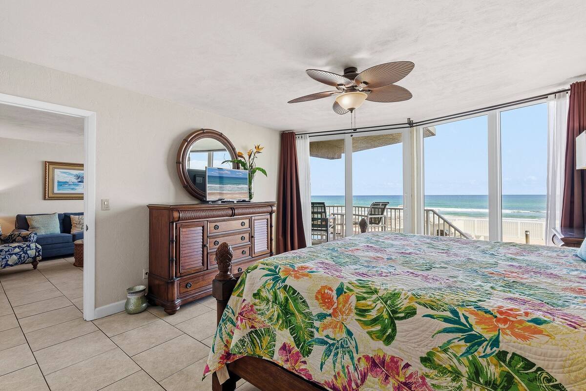 A bedroom with a large bed, a ceiling fan, a dresser, and a view of the ocean through large windows.