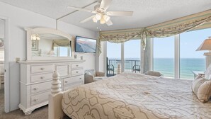 Ein Schlafzimmer mit einem großen Bett, einem Schminktisch, einem Deckenventilator, einem Fernseher und einem Balkon mit Meerblick.