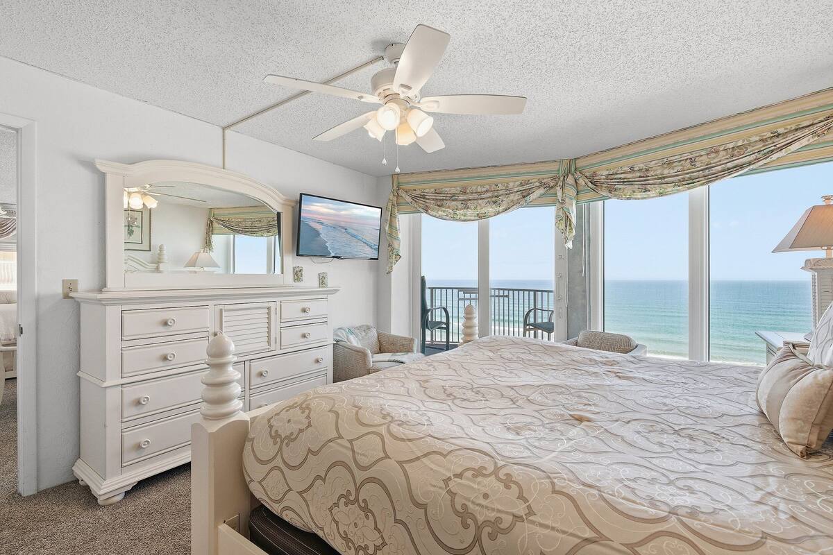 A bedroom with a large bed, a dresser, a ceiling fan, a TV, and a balcony with a view of the ocean.