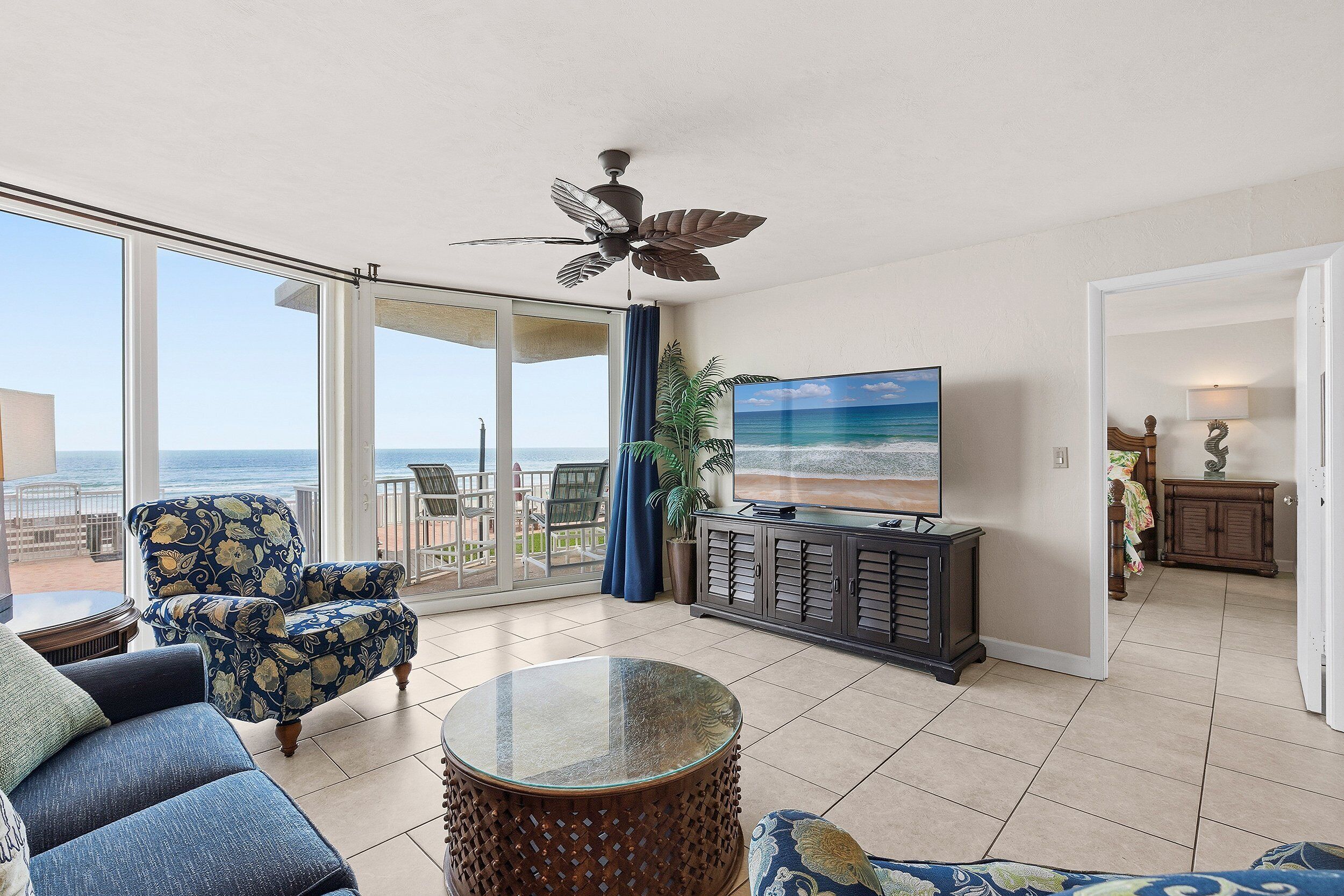 A living room with a large TV, a ceiling fan, a round coffee table, and a view of the ocean through large windows.