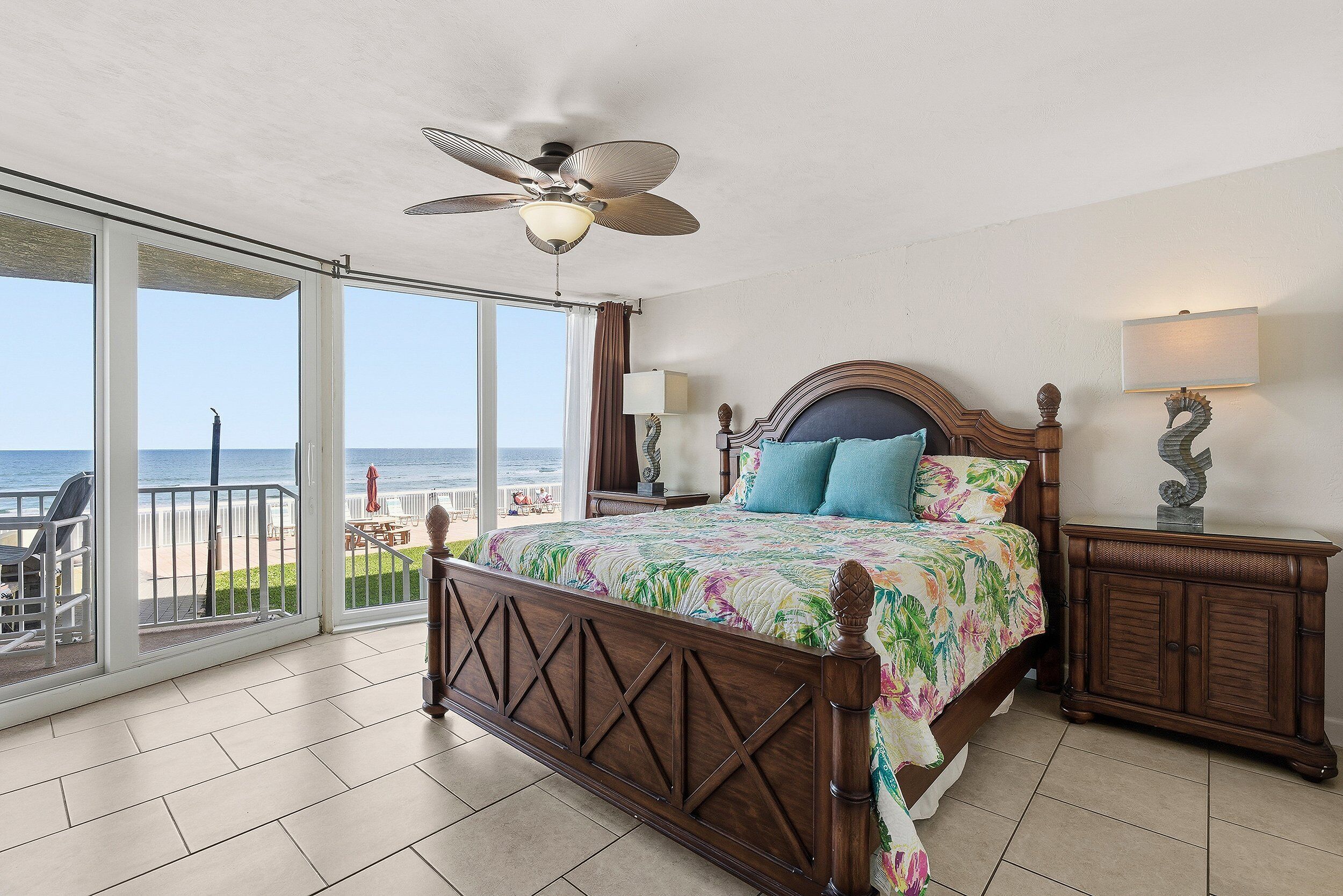 A bedroom with a bed, a nightstand, a lamp, and a ceiling fan. There is a large window with a view of the beach.