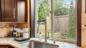 A kitchen with a granite countertop, a stainless steel sink, a blender, and a window with a view of a wooden fence and trees. - Beautiful and Pristine 4b/2.5b Home in Prime Lynnwood/Martha Lake Location (Lynnwood)