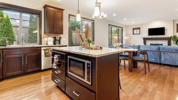 A modern kitchen with dark wood cabinets, a granite countertop, and a built-in oven. There is a dining area with a blue sofa and a TV mounted on the wall. - Beautiful and Pristine 4b/2.5b Home in Prime Lynnwood/Martha Lake Location (Lynnwood)