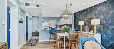 A modern living room with a dining area, featuring a chandelier, a blue sofa, and a kitchen in the background.