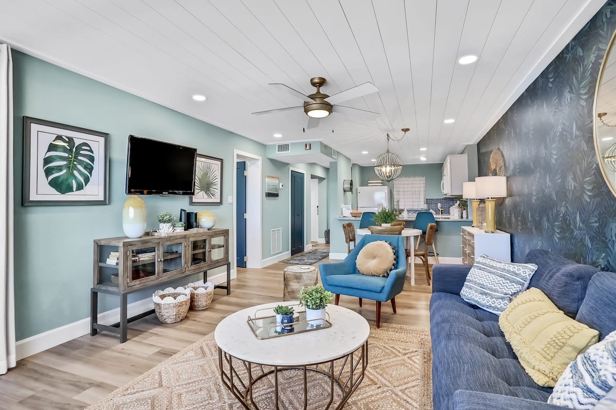 A spacious living room with a blue sofa, a coffee table, and a TV mounted on the wall.