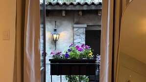 A room with a wooden floor, a bed with a dark blanket, a small table with a lamp, and a balcony with flowers and a stone wall. - Carmel loft in Val'Quirico inside Villa Alessandra (San Miguel Xoxtla)