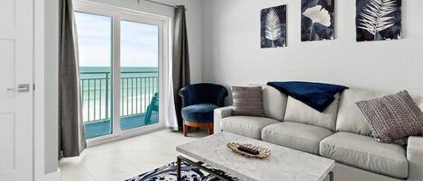 Un salon comprenant un canapé, un fauteuil et une table basse. Trois œuvres d’art ornent les murs, et de grandes fenêtres offrent une vue sur l’océan.