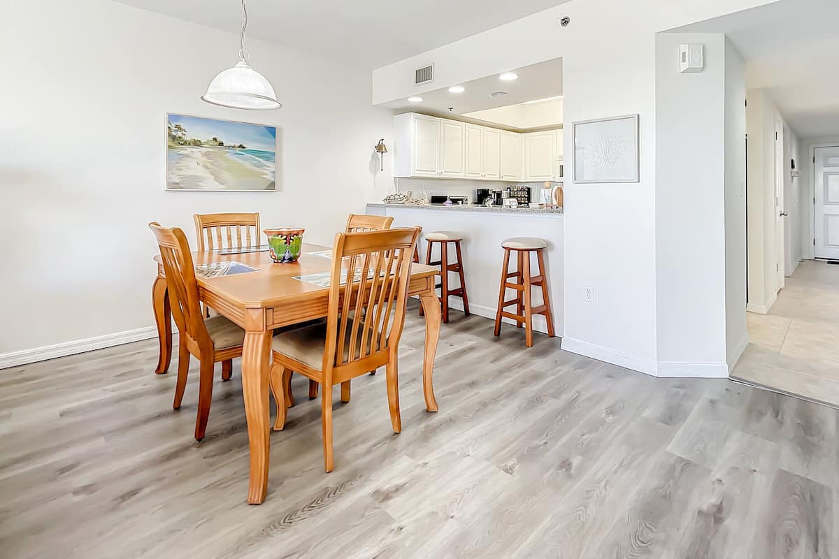 A dining area with a wooden table and chairs, a kitchen in the background, and a beach-themed painting on the wall.