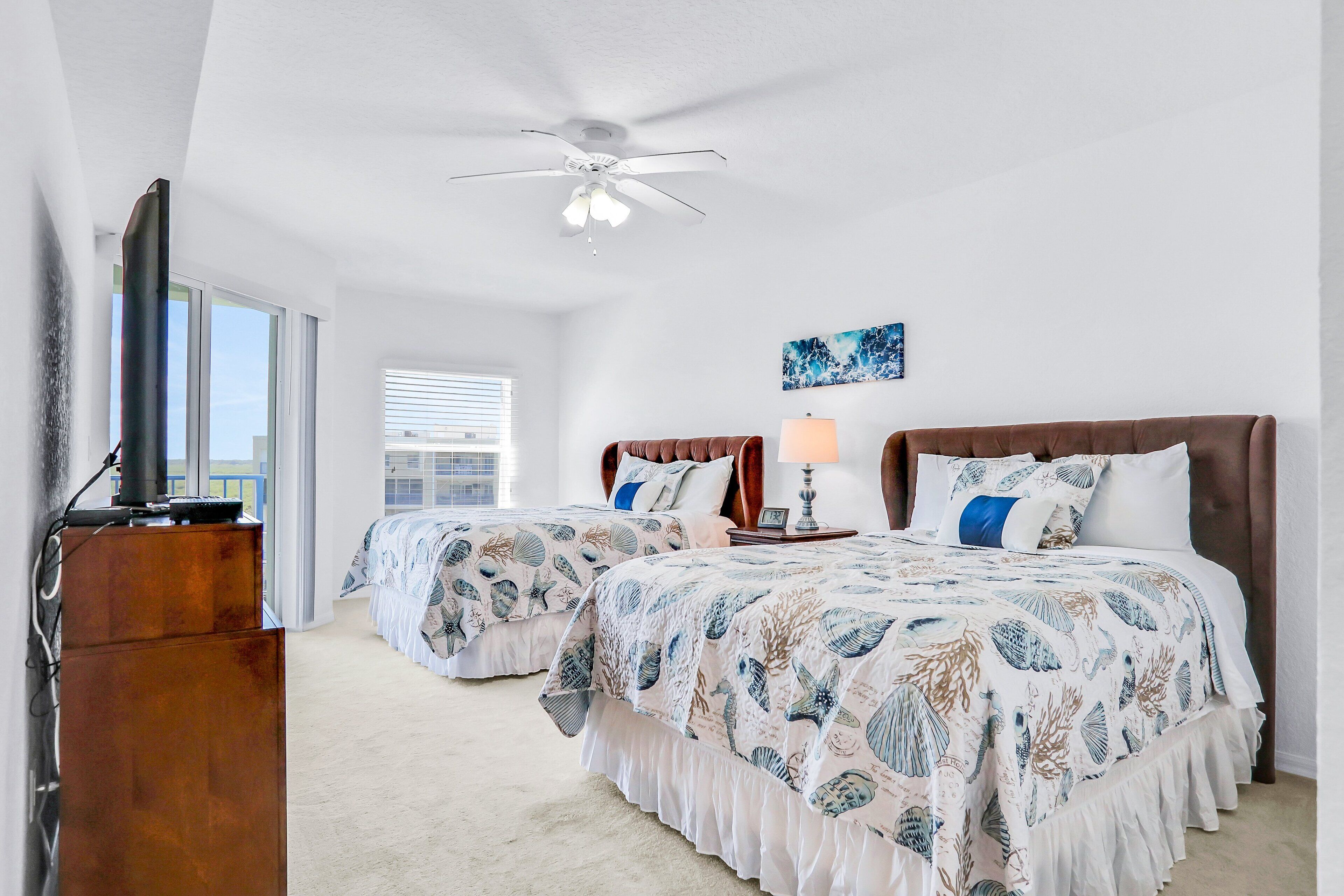 A hotel room with two beds, a ceiling fan, a TV, and a large window.