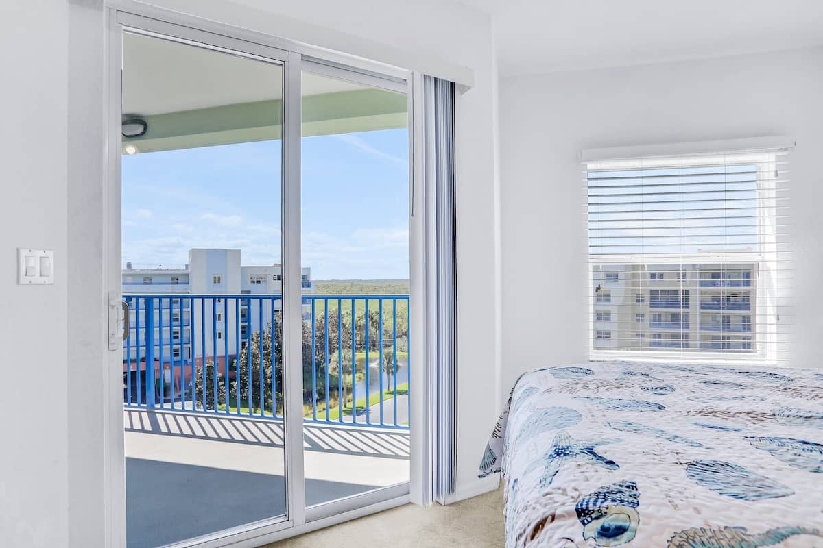 A bedroom with a large sliding glass door leading to a balcony with a blue railing, a bed with a patterned bedspread, and a window with blinds.