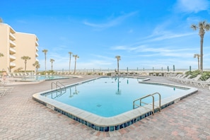 A rectangular swimming pool with a tiled edge, surrounded by lounge chairs and palm trees, with a building in the background. - Pelican (New Smyrna Beach)