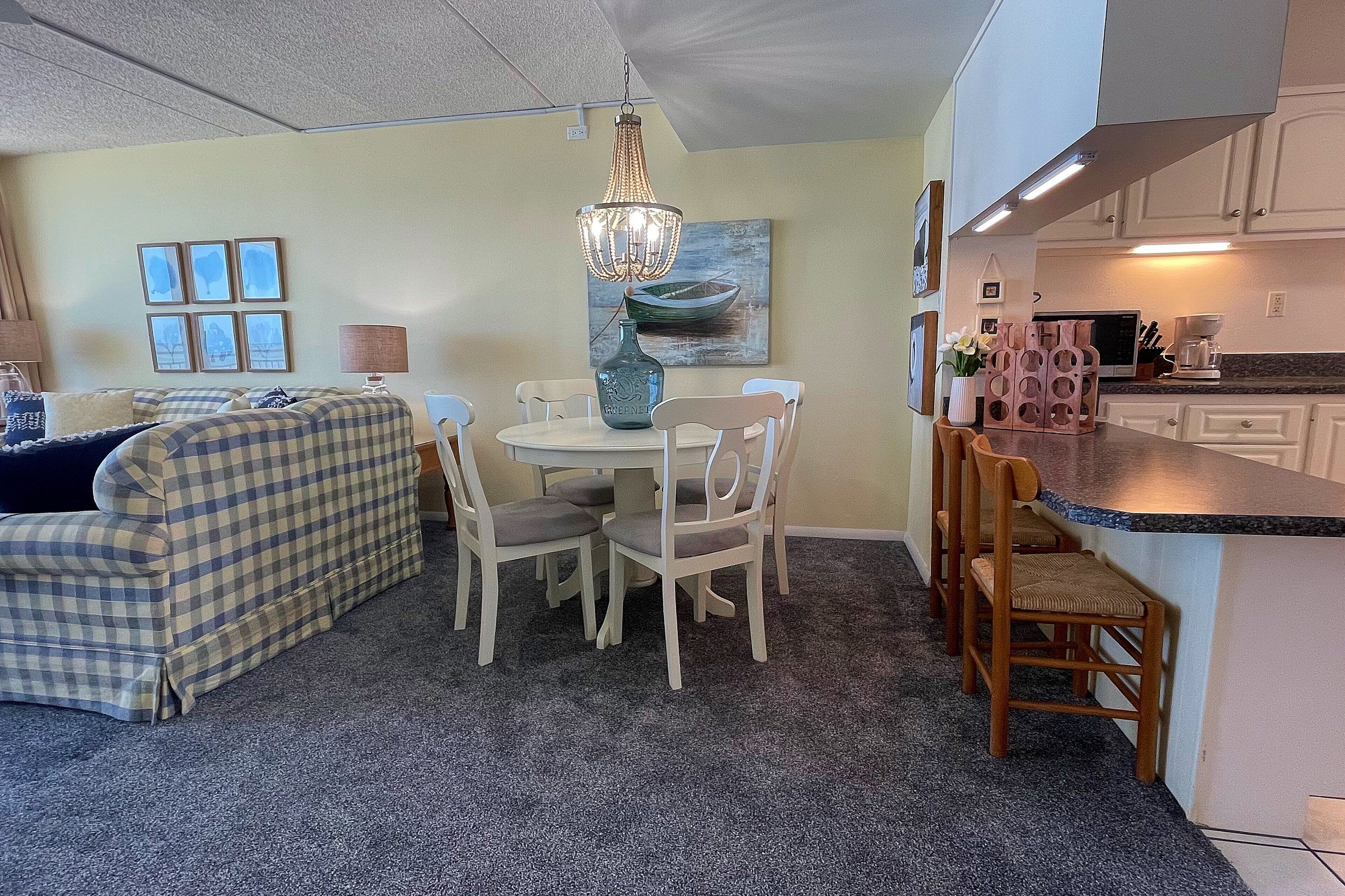 Un salon chaleureux avec un canapé à carreaux, une table à manger avec des chaises, un lustre et un coin cuisine doté d’un comptoir et de placards.
