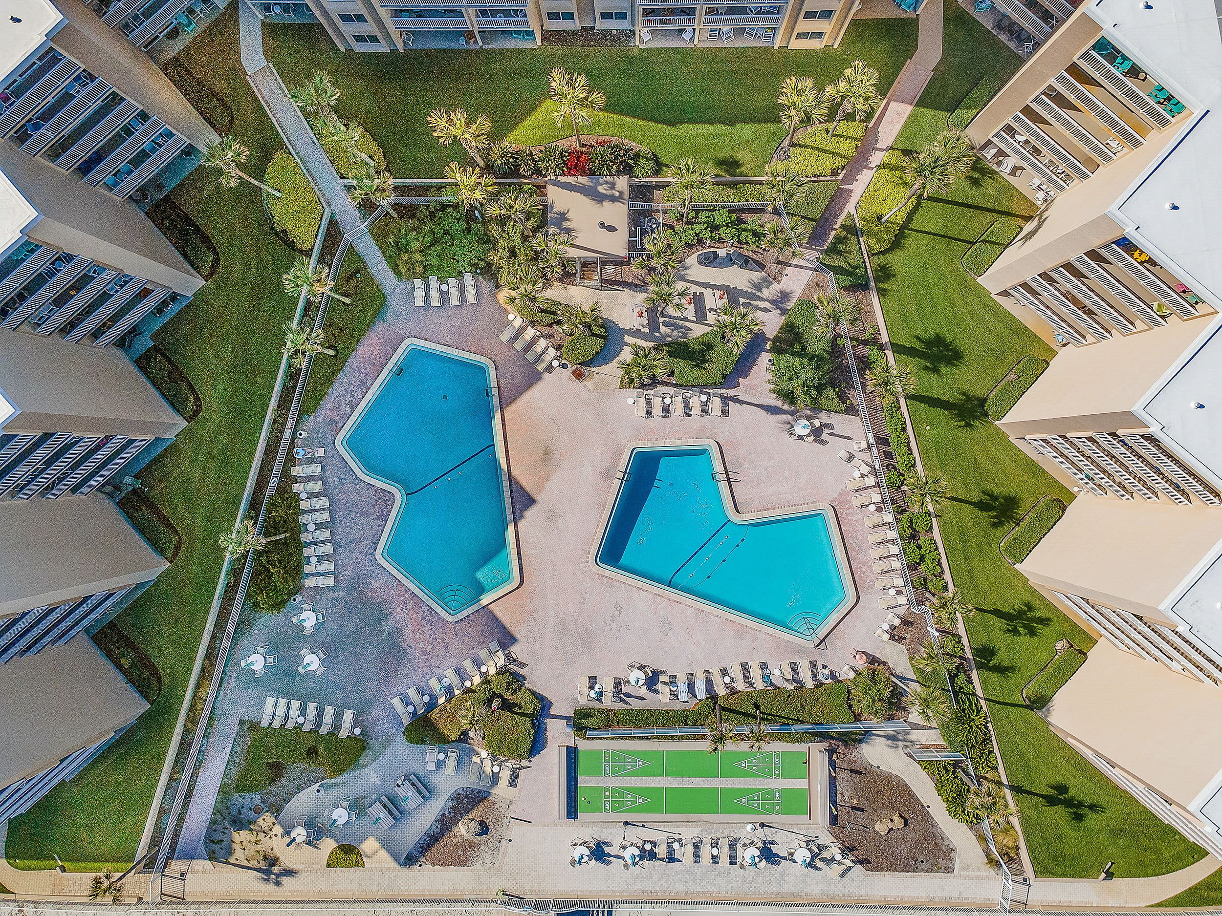 An aerial view of a hotel complex with two swimming pools, a tennis court, and a landscaped garden area.