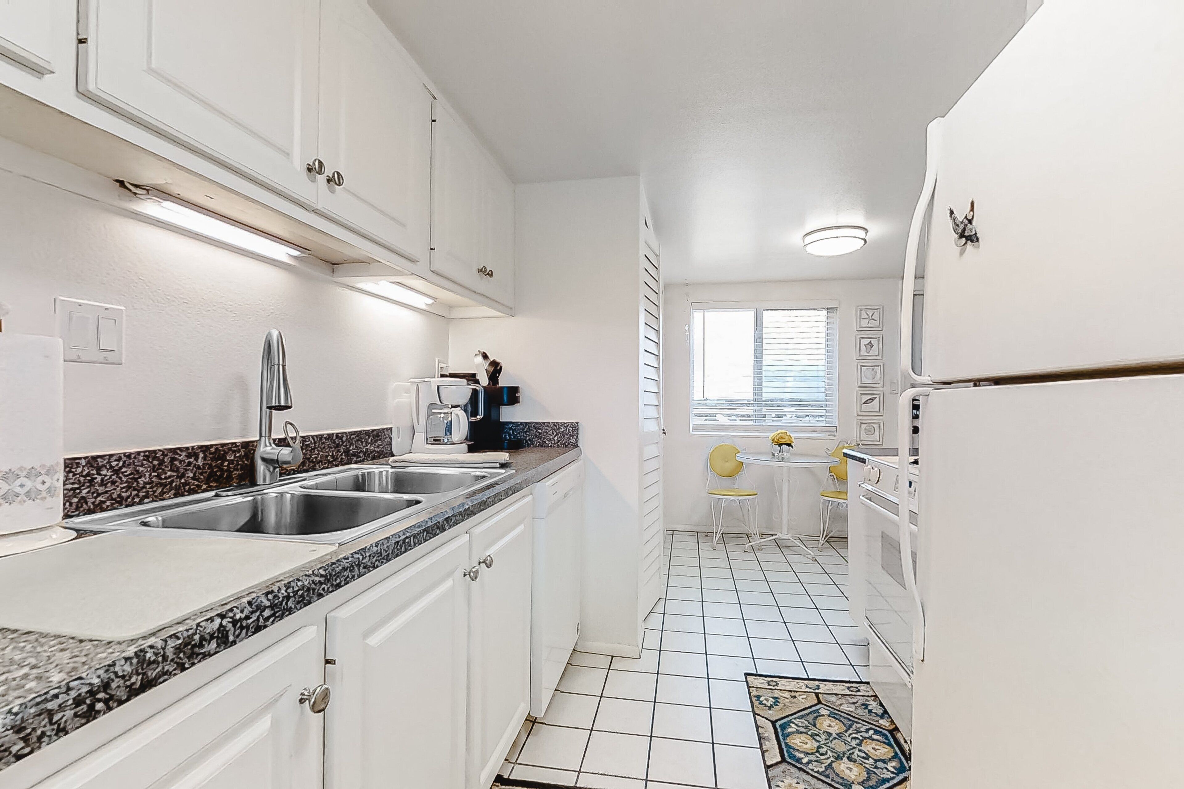 Cocina compacta con armarios blancos, encimera de granito, lavabo, cafetera y un pequeño comedor con mesa y sillas.