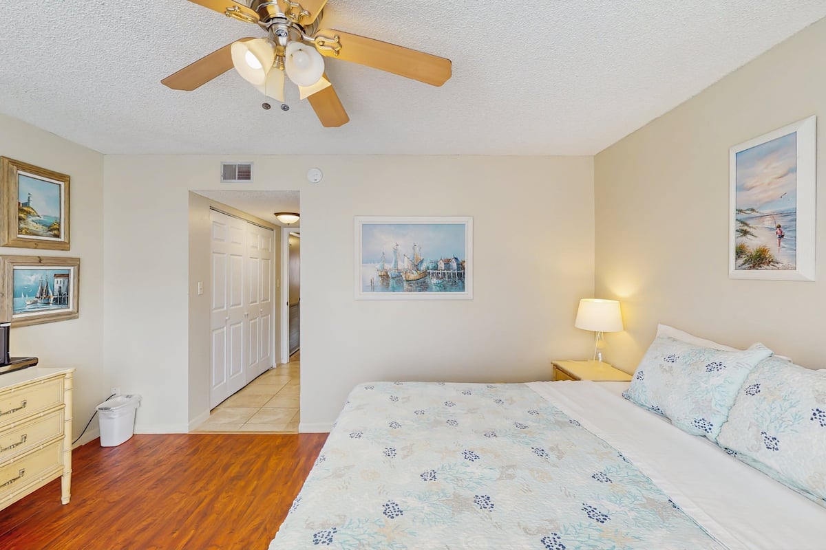 A bedroom with a bed, a ceiling fan, a nightstand with a lamp, and a door leading to another room.