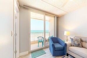 A room with a sliding glass door leading to a balcony with a view of the beach. There is a blue armchair and a beige sofa with a patterned pillow. - Ponce Inlet (Ponce Inlet)