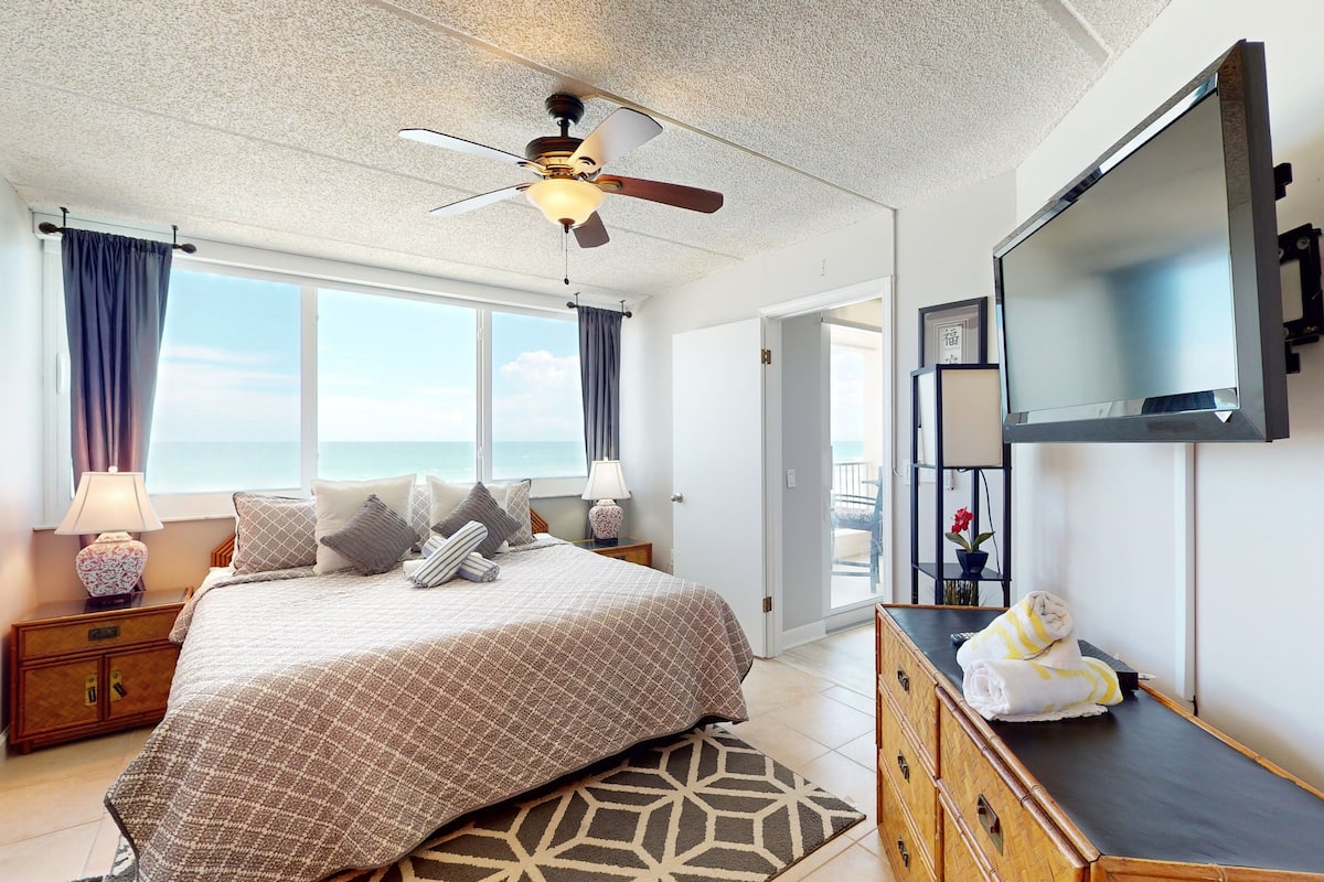 A bedroom with a bed, a ceiling fan, a TV, a dresser, and a view of the ocean.