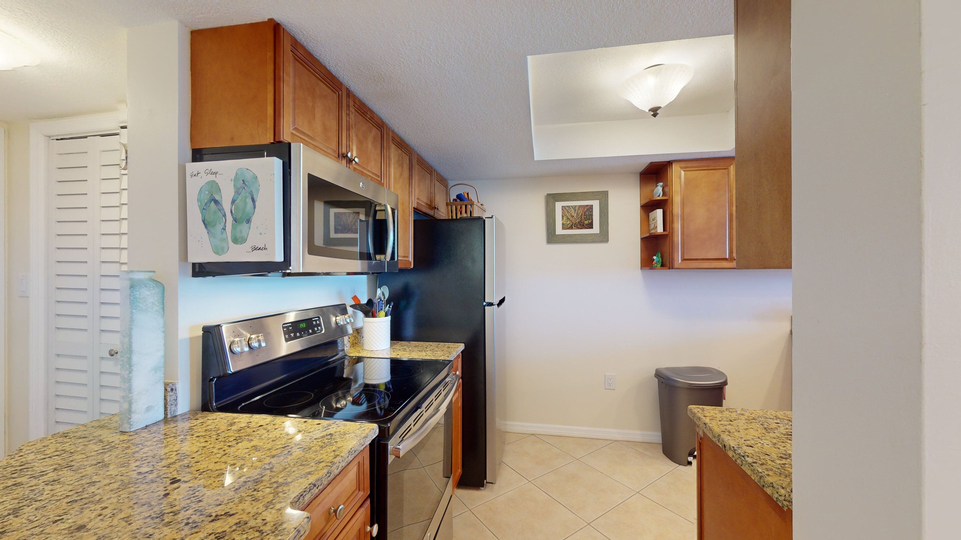 A kitchen with wooden cabinets, a stainless steel refrigerator, a microwave, an oven, granite countertops, and a trash can.