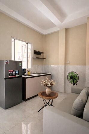 A modern kitchen with a refrigerator, sink, and a fan. There's a small table with a vase and a sofa in the living area.