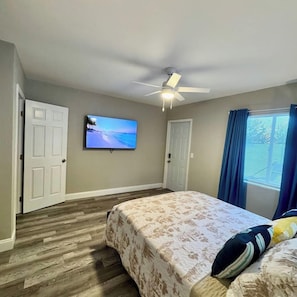 Une chambre à coucher avec un lit, un ventilateur de plafond, un téléviseur fixé au mur et une fenêtre avec des rideaux.