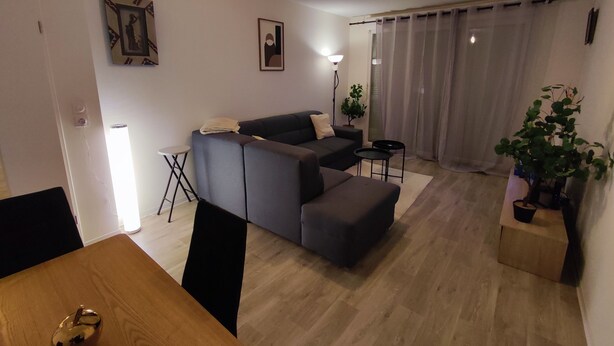 Ein modernes Wohnzimmer mit Sofa, Hocker und Esstisch. Eine Topfpflanze und eine Stehlampe sind ebenfalls vorhanden.