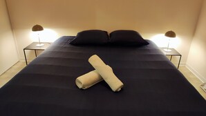 Ein ordentlich bezogenes Bett mit dunkler Bettwäsche, zwei schwarzen Kissen und einem aufgerollten Handtuch in der Mitte. Auf jeder Seite des Bettes stehen Nachttische mit Lampen.