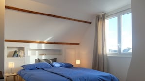 Et soveværelse med skråvægge, en seng med blåt sengetøj, et natbord med lampe, en reol med bøger og et vindue med gardiner.
