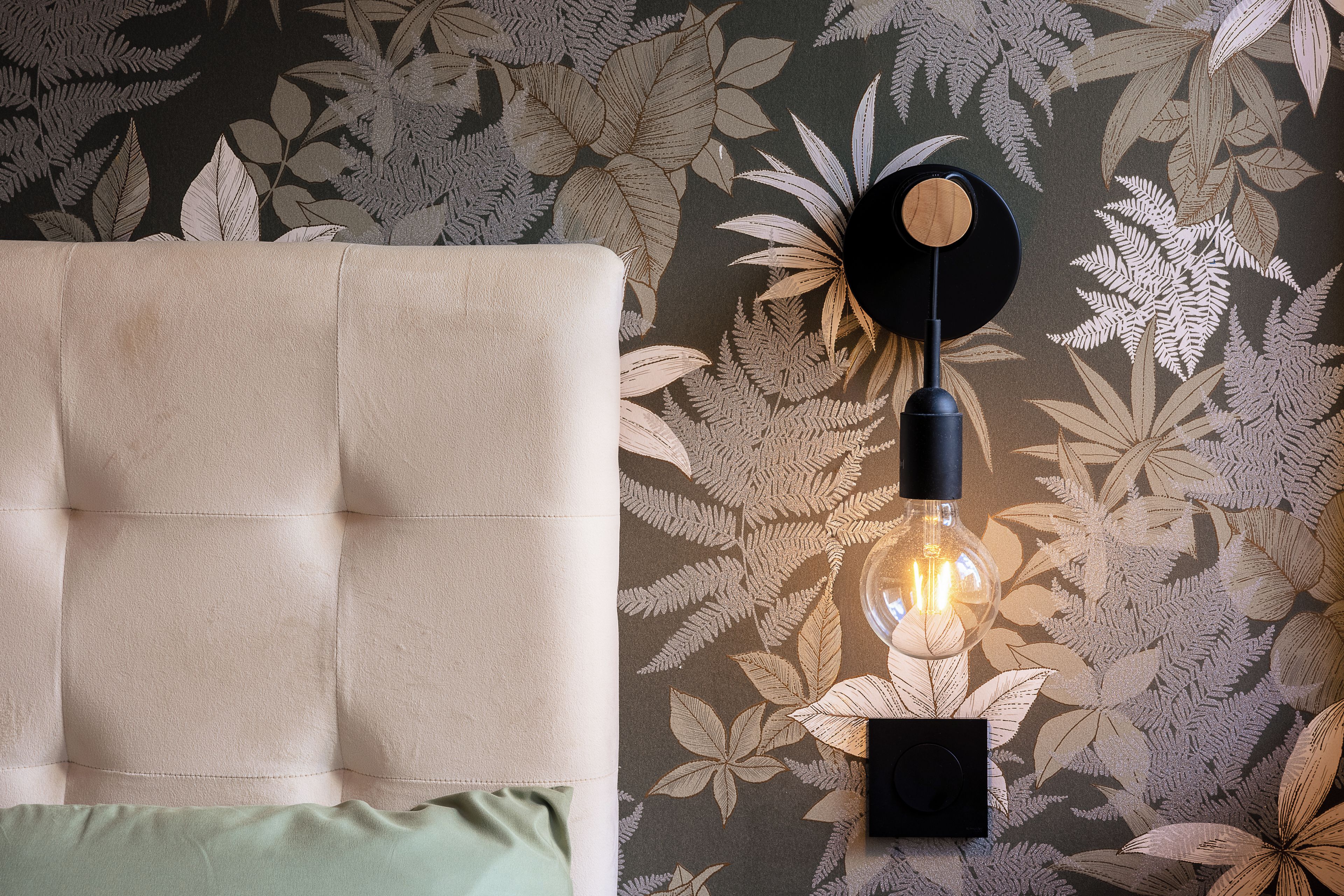 Een gestoffeerd hoofdbord met een kussen, een wandlamp met zichtbare gloeilamp, en behang met een patroon.