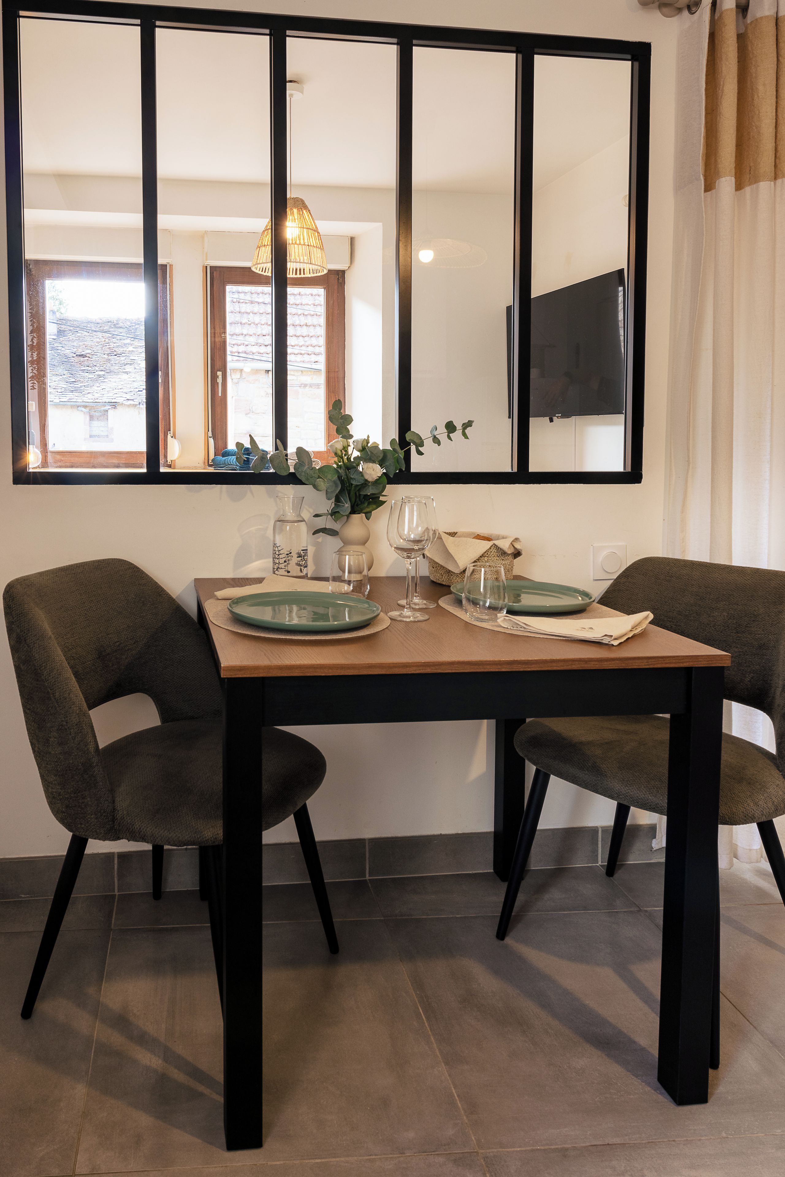 Un coin repas avec une table dressée pour deux personnes, des chaises, un vase rempli de fleurs et une fenêtre donnant sur un beau paysage.