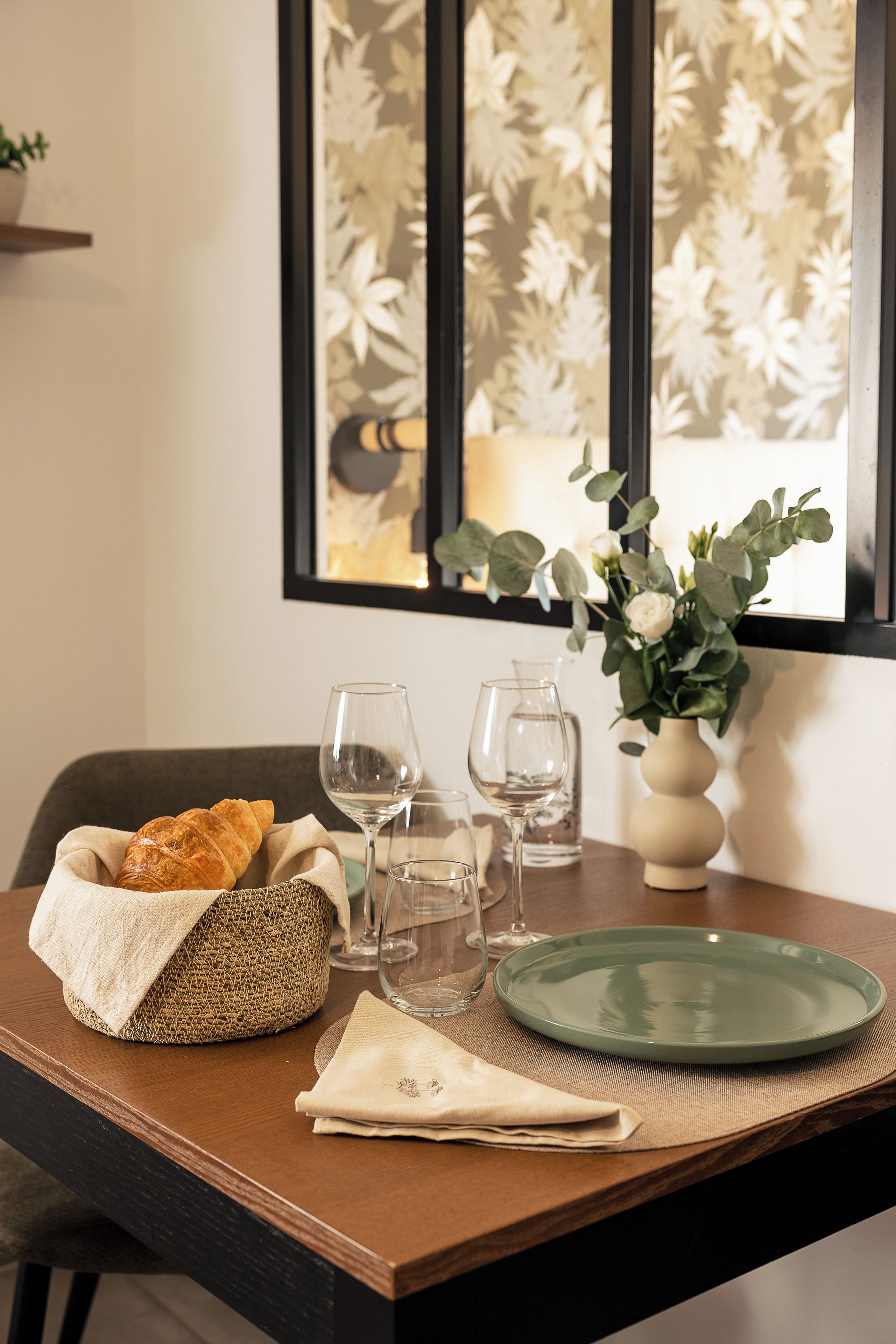 Une table à manger avec un panier tressé, une assiette verte, une serviette pliée et un vase rempli de fleurs.