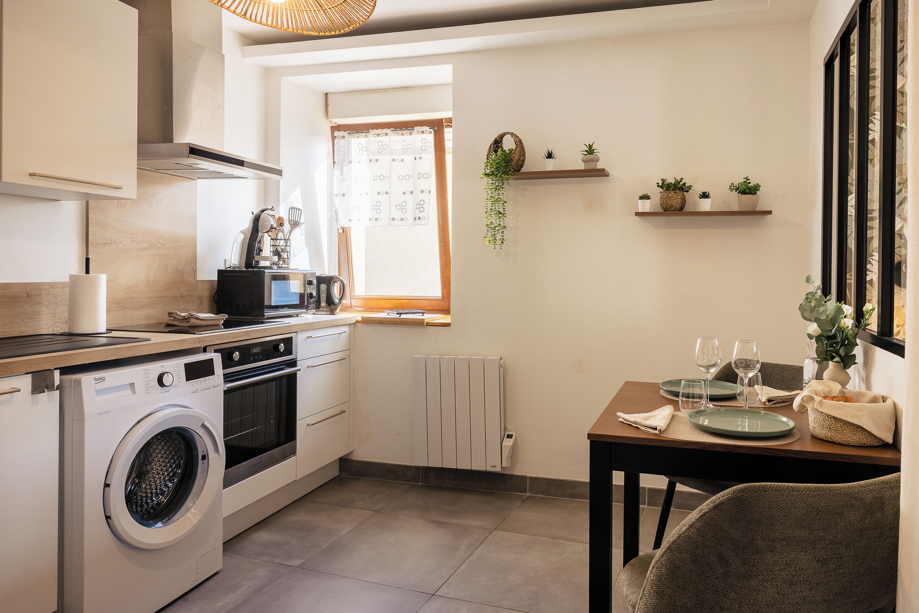 Une cuisine compacte équipée d’un lave-linge, d’une table à manger avec des assiettes et des verres, et d’une fenêtre avec des rideaux.