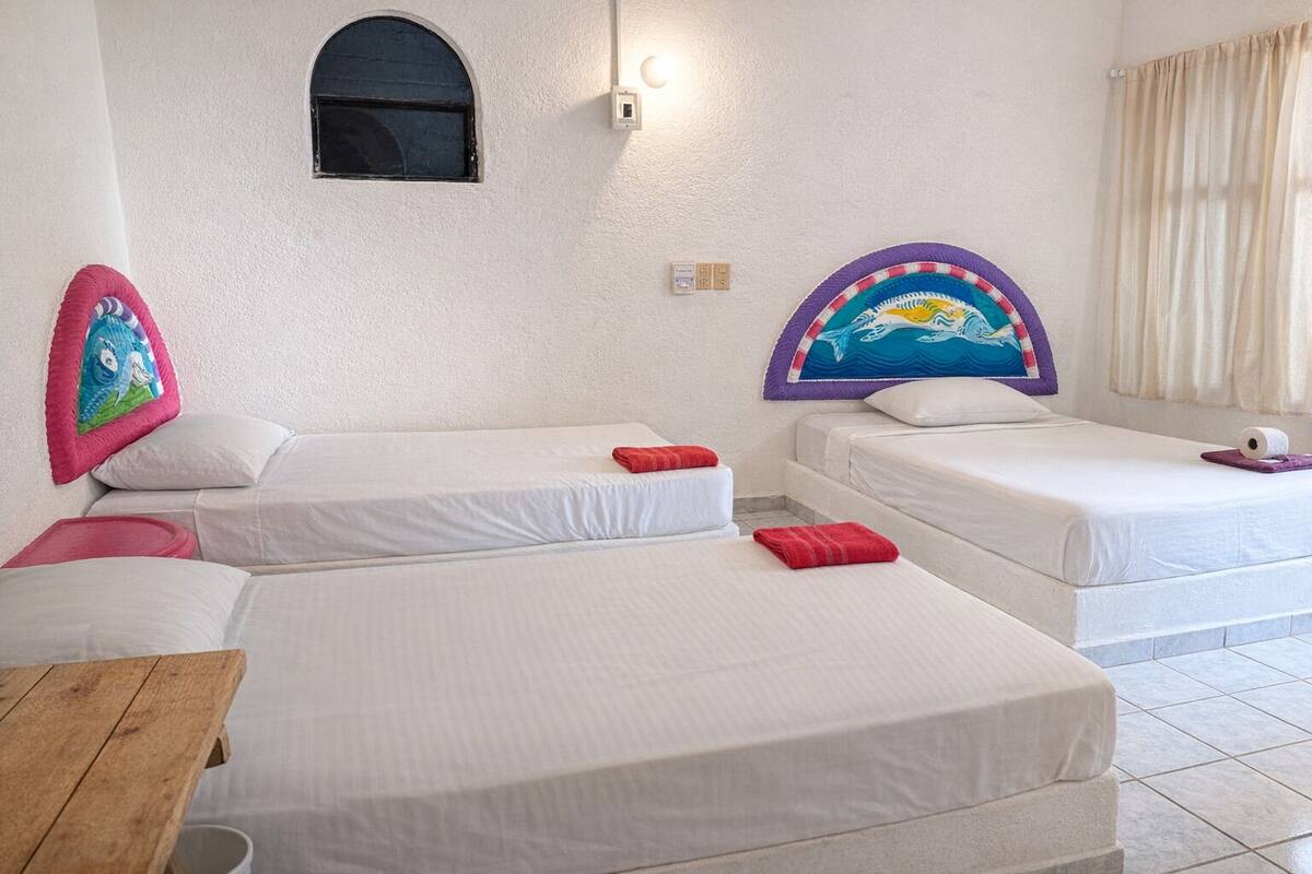 A hotel room with two single beds, a wooden table, a small window, and a wall-mounted light fixture.