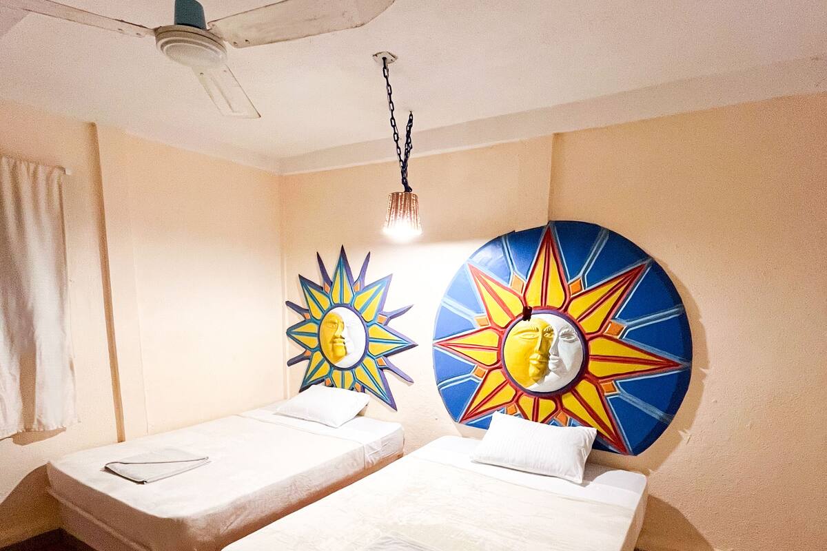 A room with two single beds, a ceiling fan, and two wall-mounted artworks.