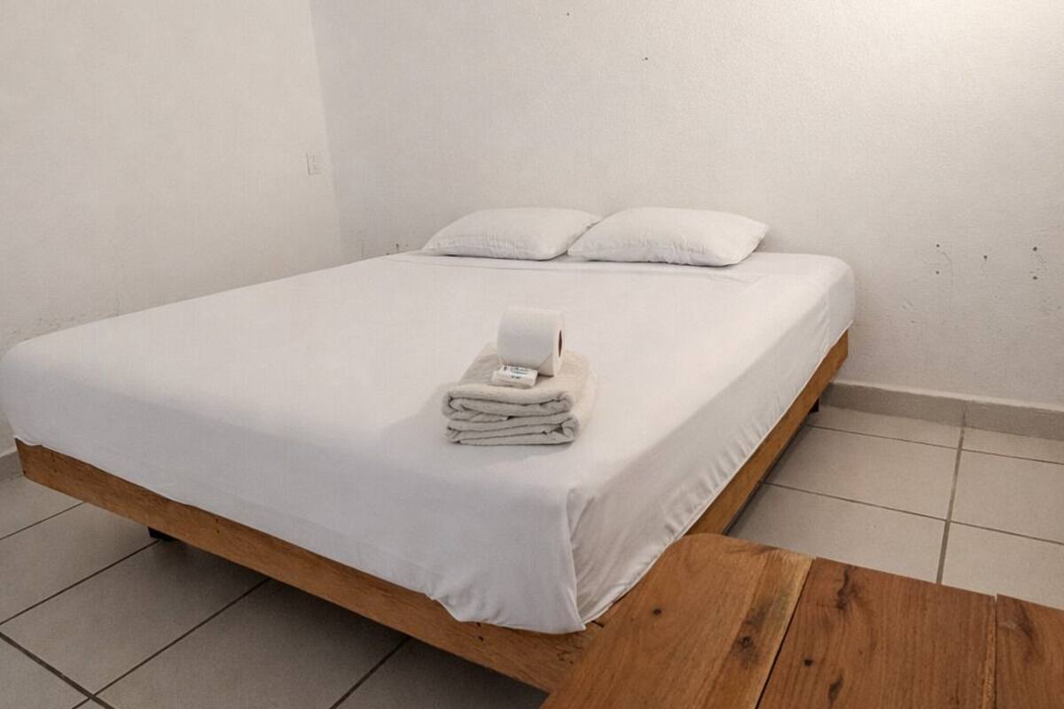 A single bed with a wooden headboard, a ceiling fan, and a wooden table in a minimalistic room with white walls and tiled flooring.