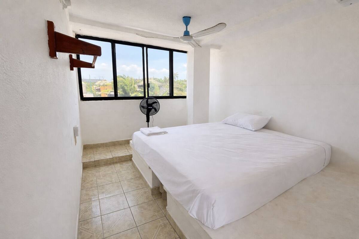 A room with a bed, a window, a ceiling fan, and a fan on the floor.