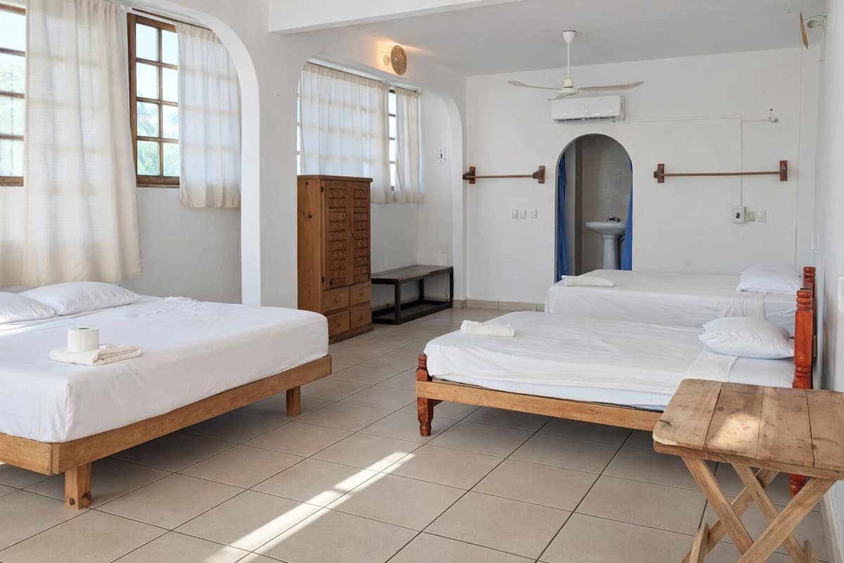 A hotel room with two beds, a wooden wardrobe, a small table, and a bathroom with a sink and mirror.