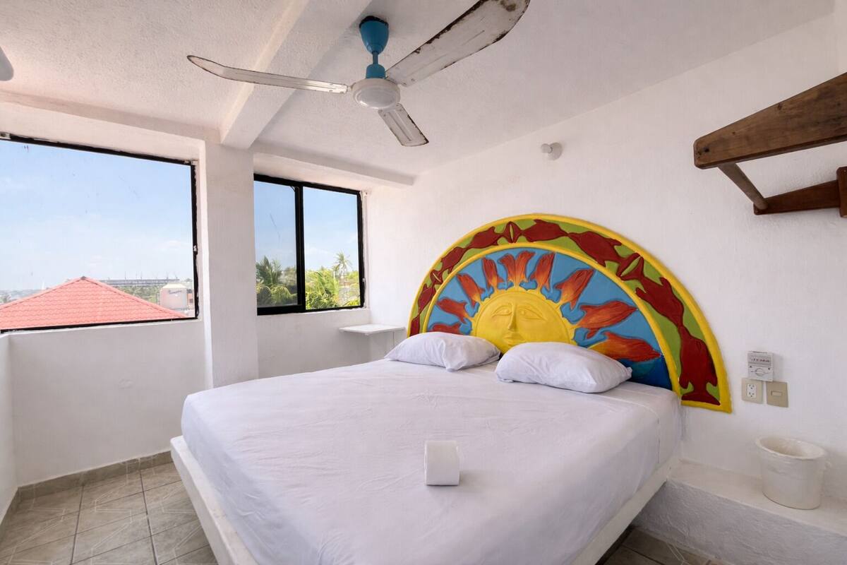 A bedroom with a bed, a ceiling fan, a window, and a view of buildings outside.